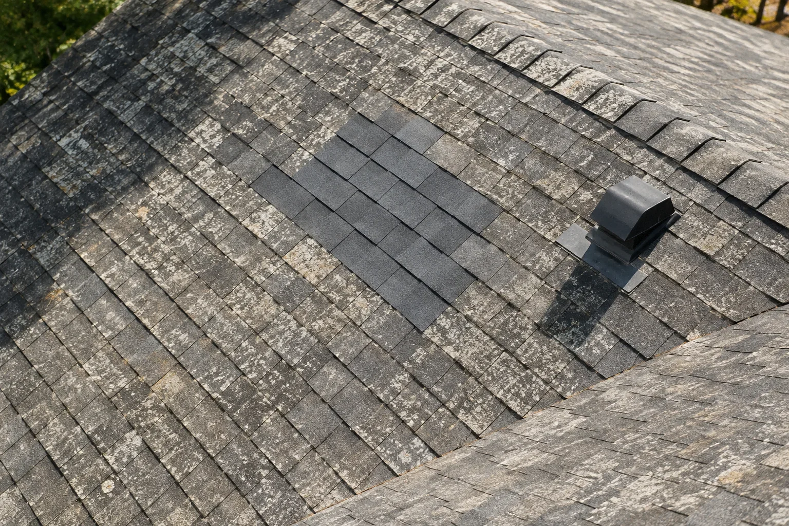 High overhead view of a residential roof with a small visibly newer repaired area amid aged covering.